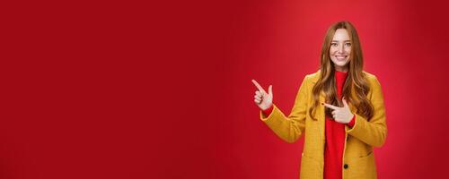 Cheerful friendly-looking and energized helpful redhead girl in yellow coat showing way pointing at upper left corner and smiling broadly with satisfied grin posing delighted over red background photo