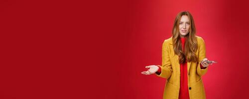 What a heck going on. Portrait of annoyed and confused perplexed ginger businesswoman in yellow coat shrugging with raised palm in clueless gesture holding smartphone troubled and questioned photo