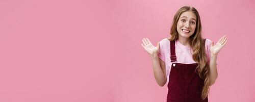 Sorry I have no idea. Silly unaware cute friendly-looking young female student in dungarees over t-shirt shrugging with palms raised in surrender smiling awkward as if being clueless over pink wall photo