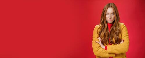 Offended and angry redhead girlfriend expressing dismay and hate as being waiting too long crossing hands over chest in defensive and aggressive pose, pouting looking with contempt over red background photo