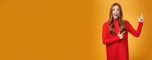 Enthusiastic and ambitious cute redhead female with freckles in red dress pointing at upper right corner smiling amazed and curious as exploring awesome new space posing over orange background photo