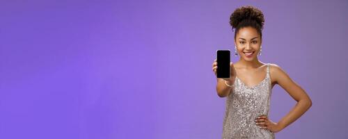 Sassy elegant african-american woman in glittering shiny silver dress hold hand waist confident pose gladly smiling extend arm showing smartphone display check out cool app blue background photo