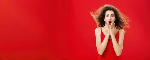 Attractive elegant young woman in red evening dress with curly hair flicking on wind saying wow being impressed and thrilled covering opened mouth from amazement standing over red background photo