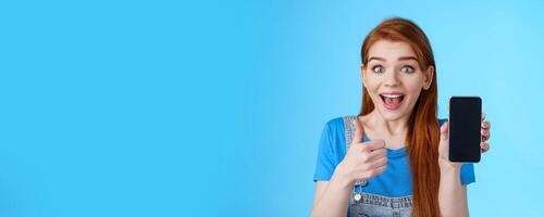 Amazed fascinated cute redhead woman recommend good smartphone, awesome new app, phone game, show cellphone display, look thrilled camera smiling approval, like cool device, blue background photo