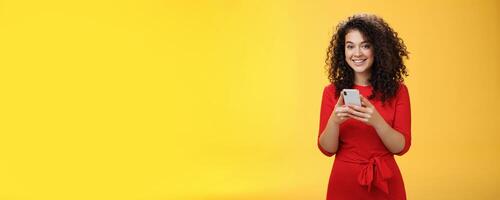 Girl messaging friend to tell all hot rumors holding mobile phone in hands smiling broadly and excited at camera as using smartphone browsing in net or edditing in app over yellow background photo