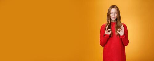Calm and confident girl showing self-assurance and professionalism as making okay gesture with unemotional expression assuring everything goes by plan standing over orange background, giving approval photo