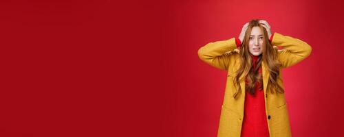 Worried and freaked out redhead girl holding hands on head grimacing troubled and concerned clenching teeth, frowning worrying and feeling nervous for project going fail over red background photo