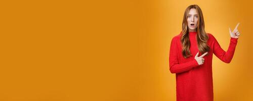 Shocked distressed and confused ginger girl with freckles and blue eyes in red sweater pointing at upper right corner with shook and surprise open mouth, raise eyebrows from stupor over orange wall photo