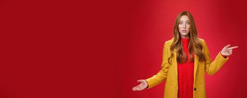 Confused and questioned cute redhead girl in yellow autumn coat pointing right as shrugging making clueless gesture with hand open mouth wondered as asking question over red background photo