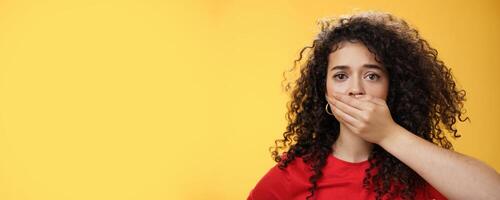 Close-up shot of concerned and insecure troubled woman keep silent scare of telling anyone her problem covering mouth with palm not to scream or slip word looking sad and sorrow over yellow background photo
