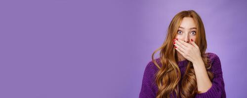 Impressed speechless cute ginger girl hearing stunning gossip covering mouth form amazement and shook raising eyebrows wondered as reacting to unexpected revelation or rumor over purple background photo