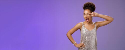 Excited lively good-looking african woman in silver shiny evening dress having fun party celebrating show lucky peace or victory gesture near eye winking happily smiling, standing blue background photo