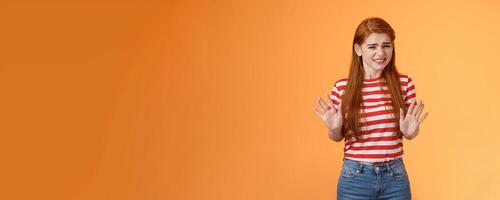Reluctant bothered cute redhead girl apologizing make stop refusal sign, hold hands block, grimacing, cringe unwilling participate, rejecting offer, stand orange background uncomfortable, step back photo
