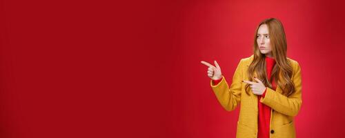 Studio shot of intense suspicious and displeased redhead female in yellow coat pointing and looking serious left squnting and frowning making focused look at something disturbing and doubtful photo