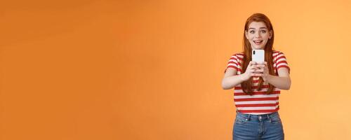 Amused excited redhead female record thrilled see famous person, smiling amazed hold smartphone taking picture, tourist adore trip shooting everything during summer journey, orange background photo