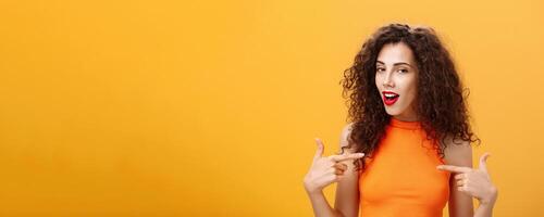 Proud and satisfied cool urban female with red lipstick and curly hairstyle pointing at herself with self-assured expression winking bragging about skills and achievements over orange background photo