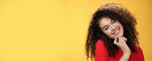 Close-up shot of charming flirty and silly curly-haired young woman making faces as trying get what wants, smiling tilting head on shoulder and touching lip standing cute over yellow background photo