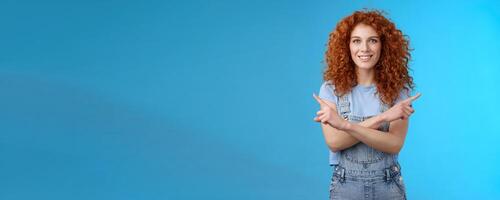 Which way. Charismatic attractive happy smiling redhead curly girl pointing curious sideways cross arms body indicating left right intrigued what choose making decision standing blue background photo