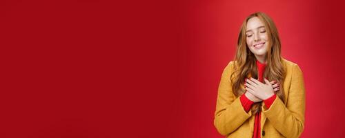Girl feeling like heart warming as remembering sweet tender memories with closed eyes and silly broad smile holding hands on chest pressing to soul posing nostalgic and touched over red background photo