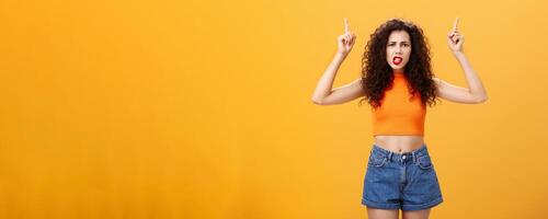 Indoor shot of displeased female client with curly stylish hairstyle red lipstick in cool cropped top pointing up demanding explanation with confused and dissatisfied expression over orange background photo