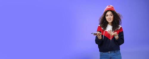 Confused and clueless girl cannot understand meaning of strange message shrugging grimacing questioned as holding smartphone and extending arm in unaware gesture over blue background photo