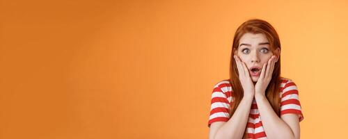 Close-up shocked speechless redhead woman, drop jaw gasping, touch cheeks embarrassed shook, look pity shame, stare camera astonished, surprised hear disturbing news, orange background photo