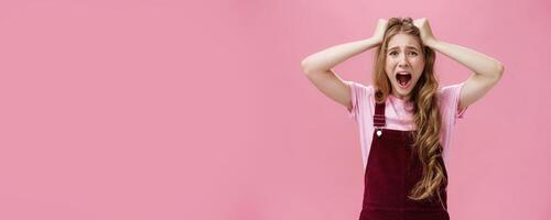Waist-up shot of woman in panic screaming and pulling hair out of head grimacing feeling anxiety from stress and problems standing bothered and distressed against pink background photo