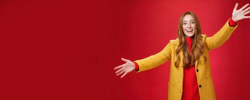 Hey come and hug me friend. Charming playful and emotional redhead female extanding palms forward to cuddle and greet, giving warm welcome smiling broadly posing in yellow coat over red wall photo