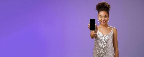 Charming attractive african-american woman in silver glittering stylish dress extend arm showing smartphone display presenting app promoting cool device standing pleased smiling blue background photo