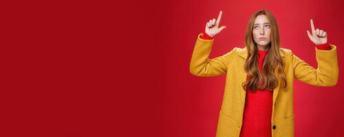 Sad and sorrow cute redhead female in yellow coat raising hands up displeased with regret looking at upper left corner apathical as being disappointed with rain or bad weather over red wall photo