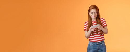 Excited redhead caucasian girl look intense, frowning, trying pass hard game level, playing new arcade, tap screen, hold smartphone horizontal, look telephone display cautious, orange background photo