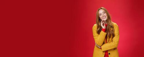 Good-looking carefree and happy relaxed redhead girlfriend in yellow stylish coat touching face and tilting head as smiling with positive emotions, posing cheerful against red background photo