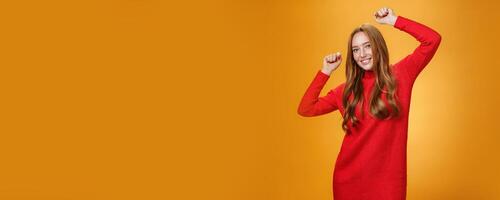 Joyful and carefree relaxed ginger girl in red warm dress partying having fun enjoying cool music as dancing with raised hands smiling broadly at camera against orange background photo