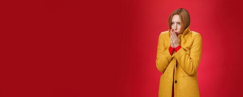 Studio shot of intense trembling cute redhead woman wearing yellow light coat rub hands and blowing warm breath at palms to warm-up feeling cold, freezing during cold windy weather over red background photo