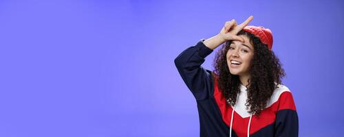 Girl triumphing and mocking friend as winning showing loser gesture on forehead and laughing having fun standing pleased and happy over blue background, smiling at camera joyfully photo