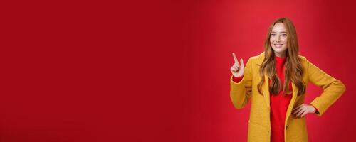 Charming young female in yellow coat holding hand on waist and pointing at upper left corner, smiling broadly as showing or promoting interesting proposal over red background, in good mood photo