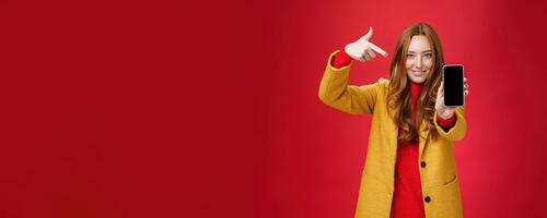 Pick this phone you never regret. Portrait of friendly-looking attractive and confident redhead glamour girlfriend in yellow coat showing smartphone pointing at mobile as smiling broadly at camera photo