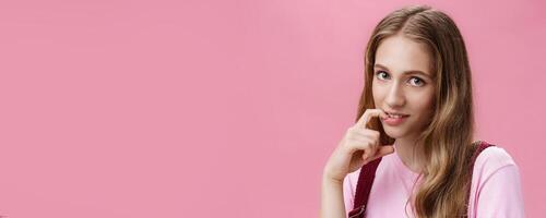 Cute feminine girl learning how make glances and seduce boyfriend with flirty gaze biting finger sensually posing against pink background, flirting making senducing look wearing make-up on date photo