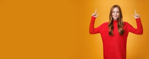 Look up you not regret it. Portrait of confident and stylish young redhead woman in 20s wearing knitted red dress pointing upwards and smiling assertive with self-assured grin over orange background photo