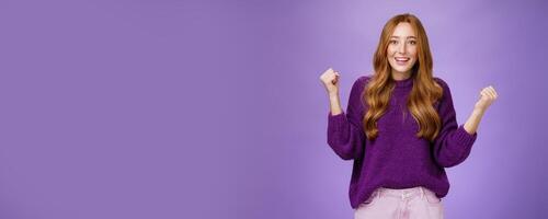 Girl proud of herself making successful effort clenching raised fists in celebration and triumph smiling broadly with surprised and relieved expression, winning, achieving goal over purple background photo