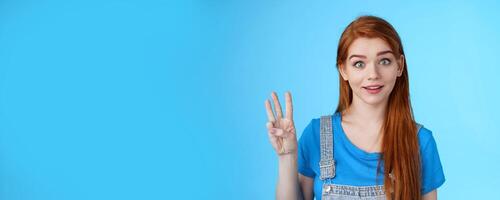 Surprised cute redhead caucasian girl show number three, third, counting fingers, raise eyebrows ambushed, smiling curiously, stand blue background playful tender expression photo