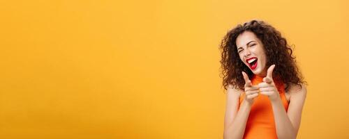 Hey what is up. Friendly and outgoing attractive curly-haired female with red lipstick pointing at camera with finger guns as if greeting or hinting friend winking at camera over orange background photo