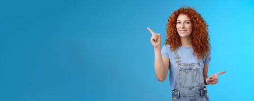 Lively joyful good-looking 25s redhead curly-haired sassy girl summer positive mood dancing overalls pointing sideways up right index fingers directing promos store links smiling delighted photo