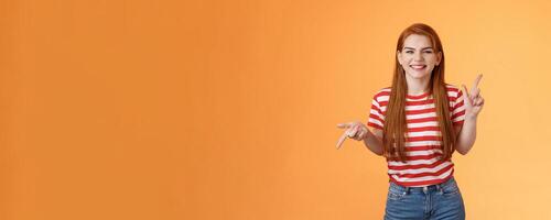 Carefree daring redhead cheerful girl dancing, pointing sideways, showing different sides, left right copy space, laughing and smiling broadly, joyfully recommend cool product, orange background photo