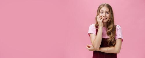 Woman panicking and overreacting feeling nervous biting fingernails and staring afraid at camera trembling from fear over pink background posing in cute dungarees with natural wavy hairstyle photo