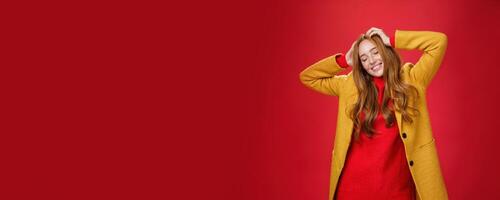 Carefree happy ginger girl carrying away with music and good vibes holding hands on head close eyes and smiling delighted and relaxed having fun, dancing against red background in yellow coat photo