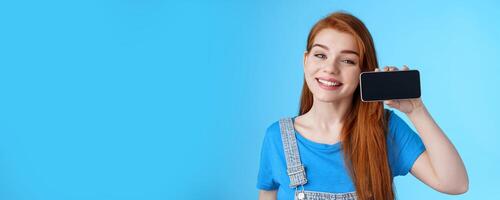 Close-up cheerful amazed cute redhead female look delighted, hold smartphone horizontal, show application, favorite phone game, stand blue background satisfied happy, grinning pleased photo