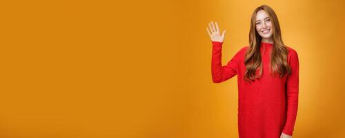 Friendly and optimistic good-looking ginger girl in red sweater raising palm waving at camera, saying hello or hi joyfully smiling cute, greeting new members posing over orange background photo