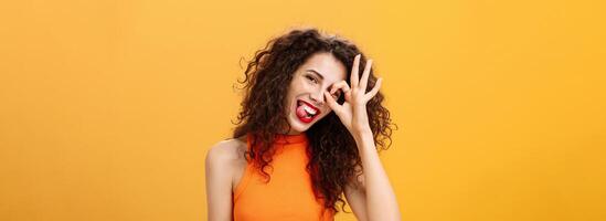 Carefree optimistic and energized good-looking woman with curly hairstyle tilting head and sticking out tongue playfully showing okay or excellent sign over eye posing near orange background photo