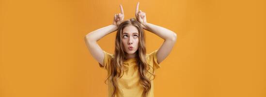 GIrl going crazy from boredom making freaky funny faces fooling around squinting holding index fingers on head like horns sticking out tongue and folding lips aping over orange background photo